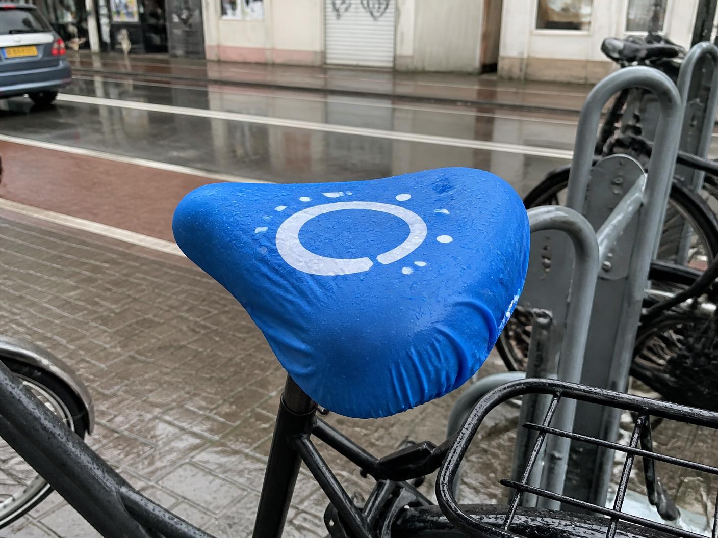 Blauw bedrukt zadelhoesje met logo op een stadsfiets in een natte Nederlandse straat
